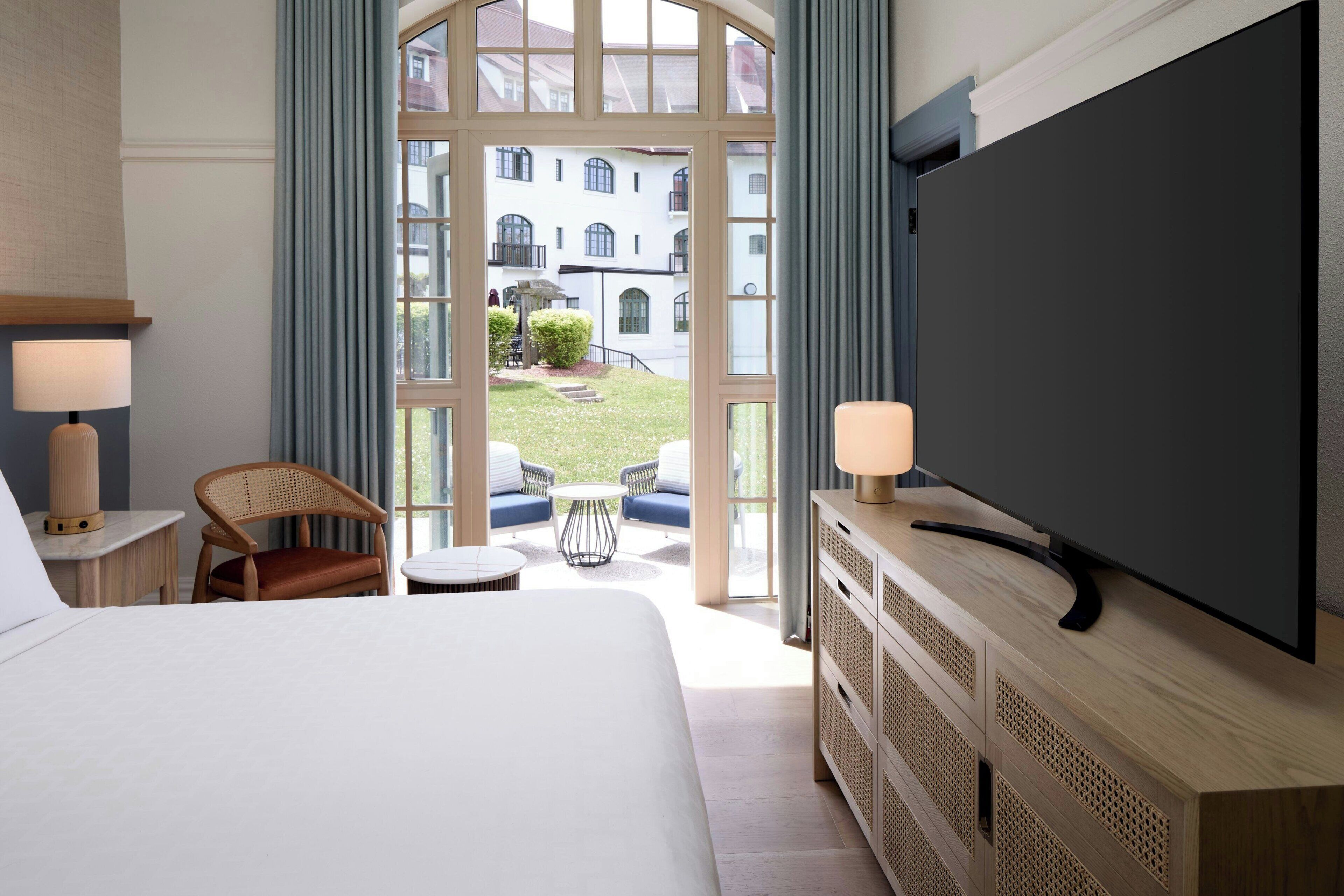 Quarto de hotel com uma cama grande, uma televisão sobre um armário de madeira, uma cadeira, uma mesa pequena e vista para um pátio com edifícios.