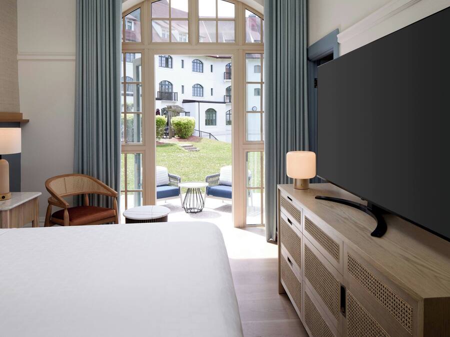 Quarto de hotel com uma cama grande, uma televisão sobre um armário de madeira, uma cadeira, uma mesa pequena e vista para um pátio com edifícios.