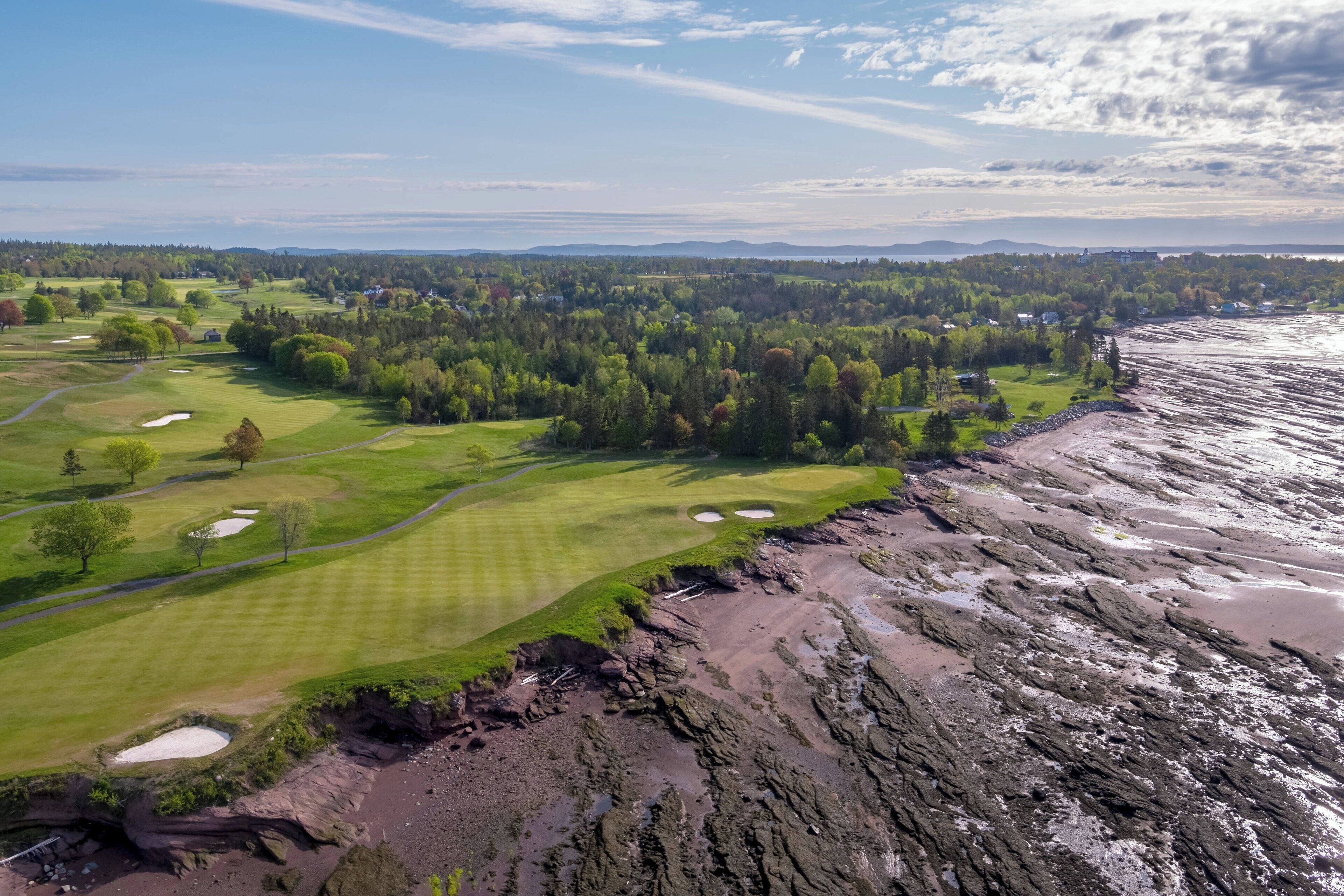 A coastal golf course with a rocky shoreline, lush greenery, and distant mountains.