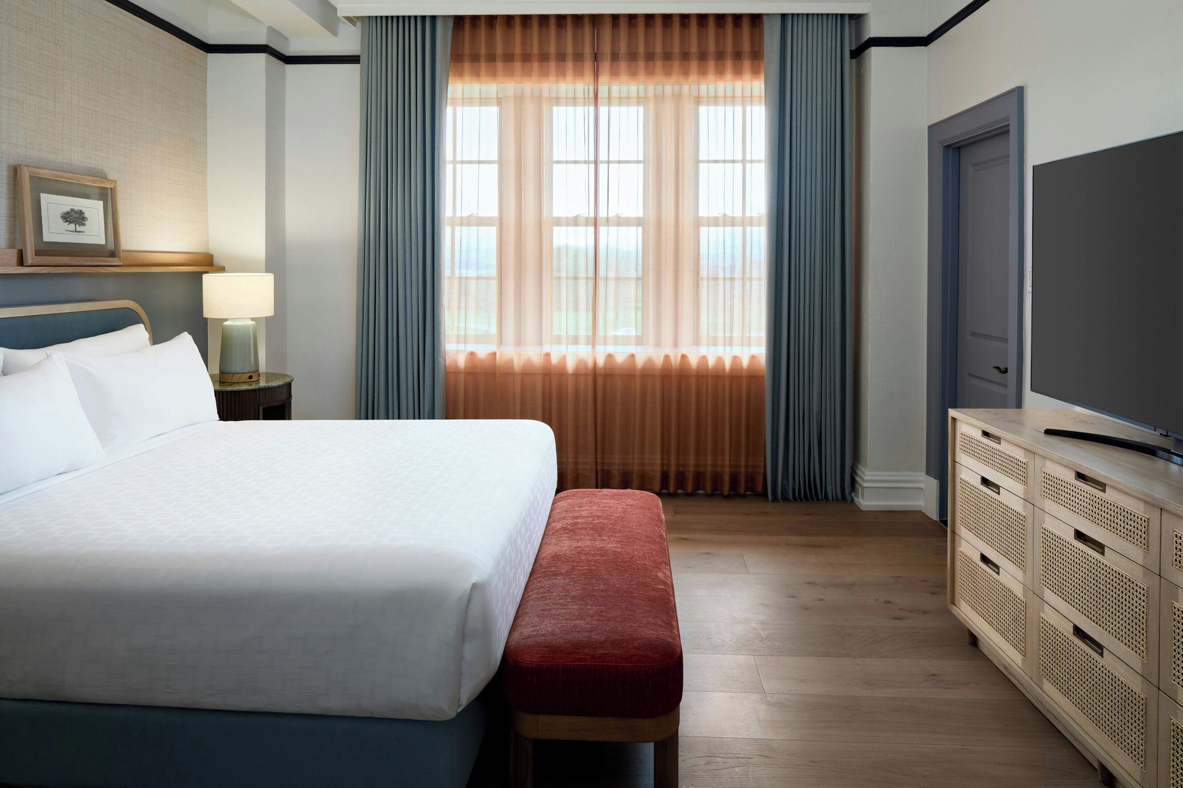 A hotel room with a bed, a bench, a TV, a nightstand, a lamp, and a window with curtains.