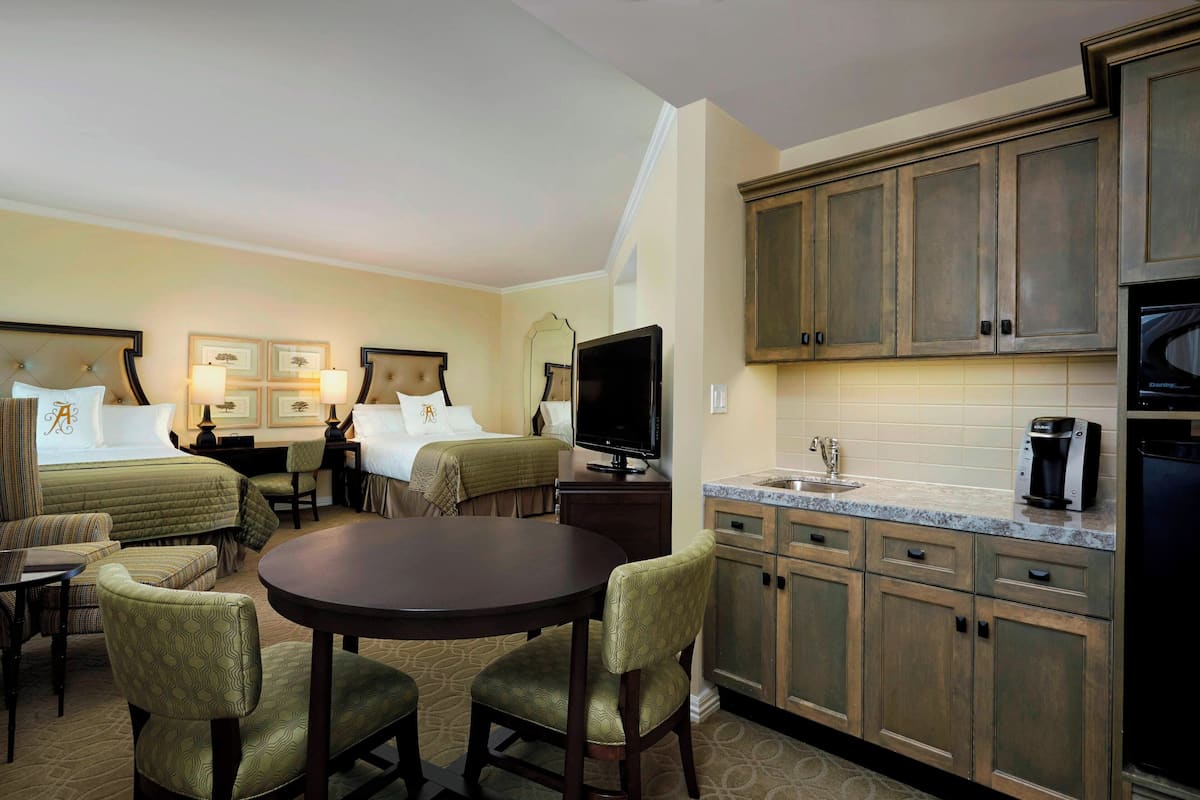 Une chambre d’hôtel comprenant un lit, deux tables de chevet, un téléviseur, une table à manger avec des chaises, ainsi qu’un coin cuisine avec des placards et un four à micro-ondes.