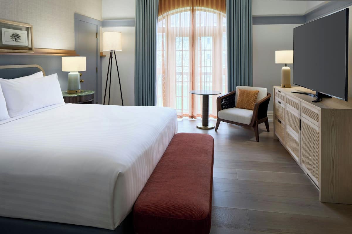 Une chambre d’hôtel avec un grand lit, une chaise, une petite table, un téléviseur et une fenêtre avec des rideaux.