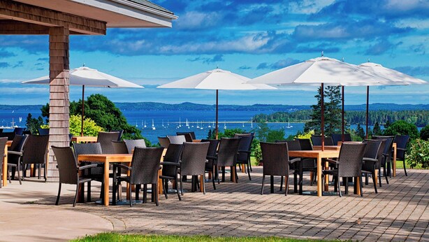 Un patio al aire libre con mesas y sillas, sombrillas grandes y una vista panorámica del agua y las montañas.