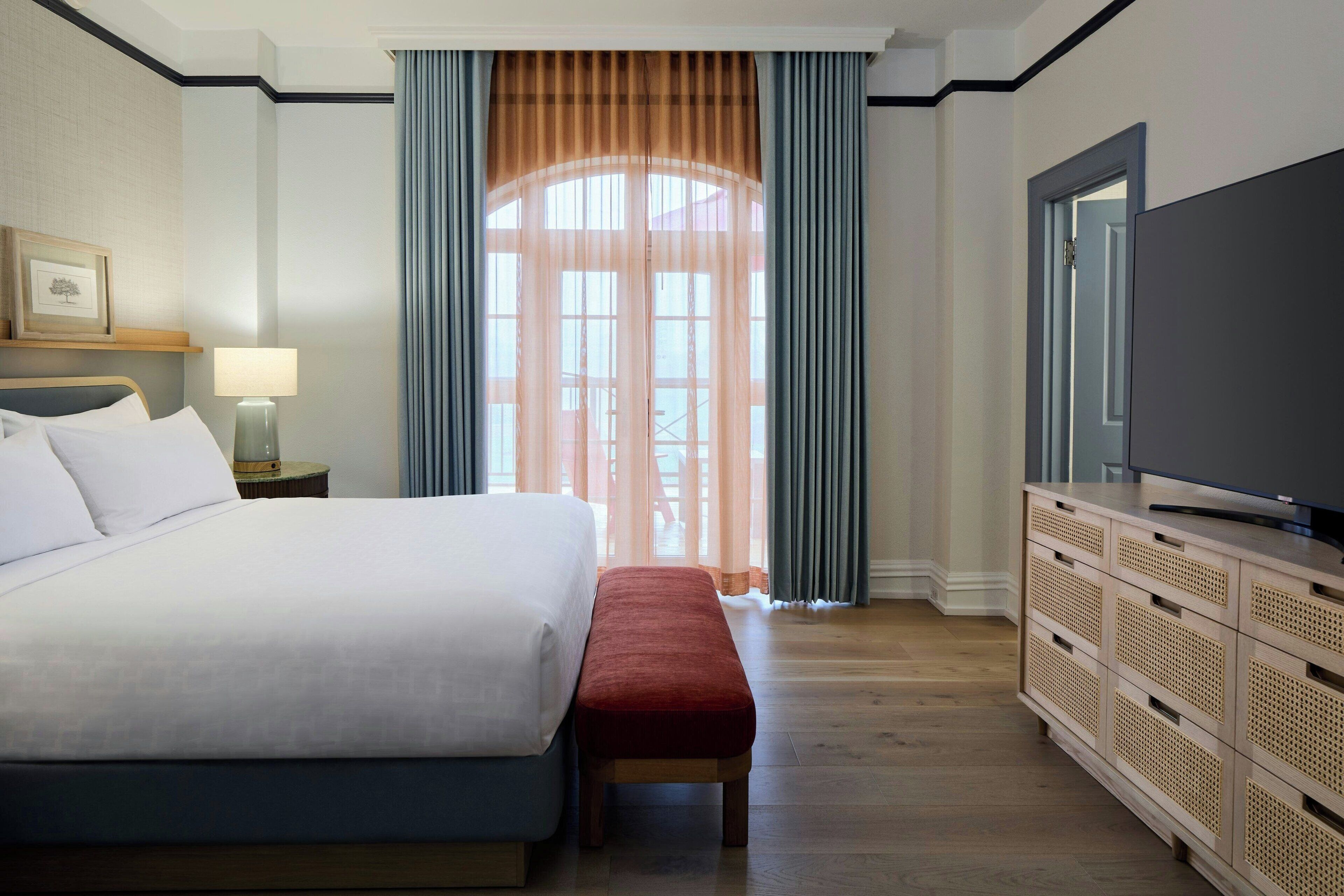 Habitación de hotel con una cama grande, un banco, un televisor sobre un mueble de madera y una ventana con cortinas.