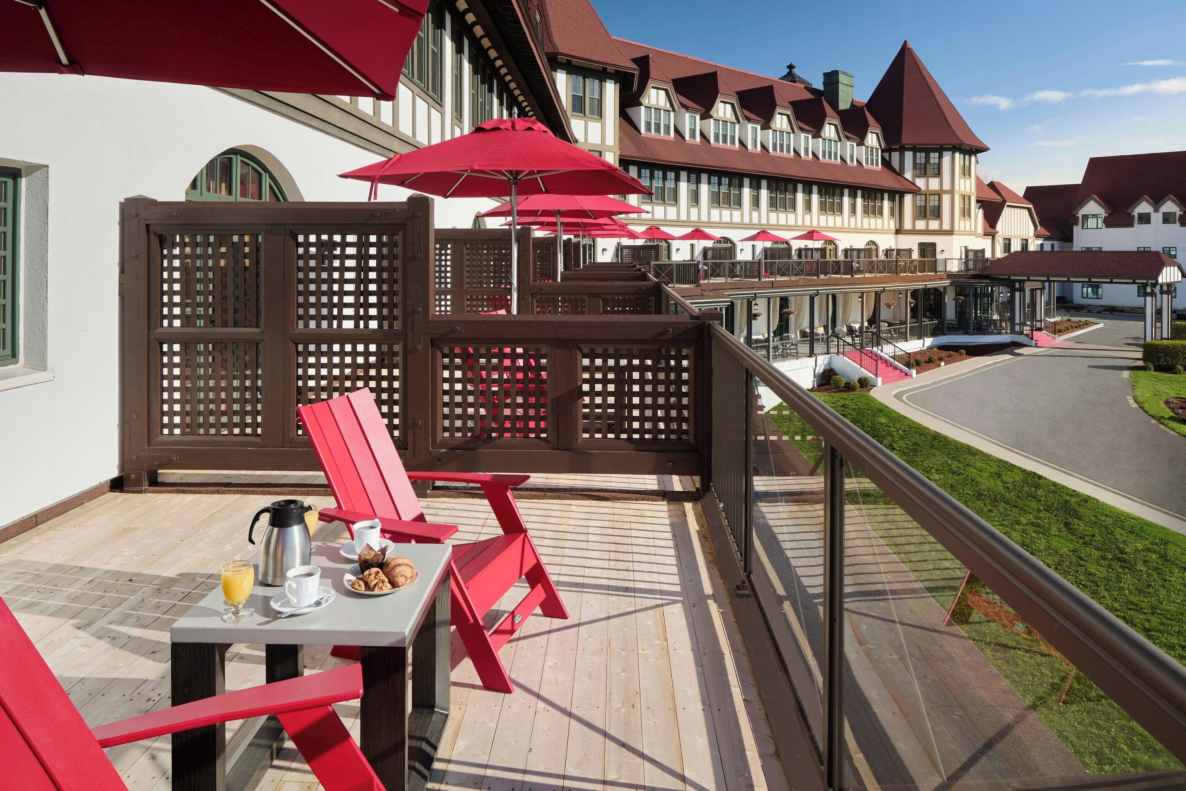 Un balcón con sillas y sombrillas rojas, una mesa con alimentos para el desayuno, y vistas a un edificio con múltiples ventanas y techo rojo.