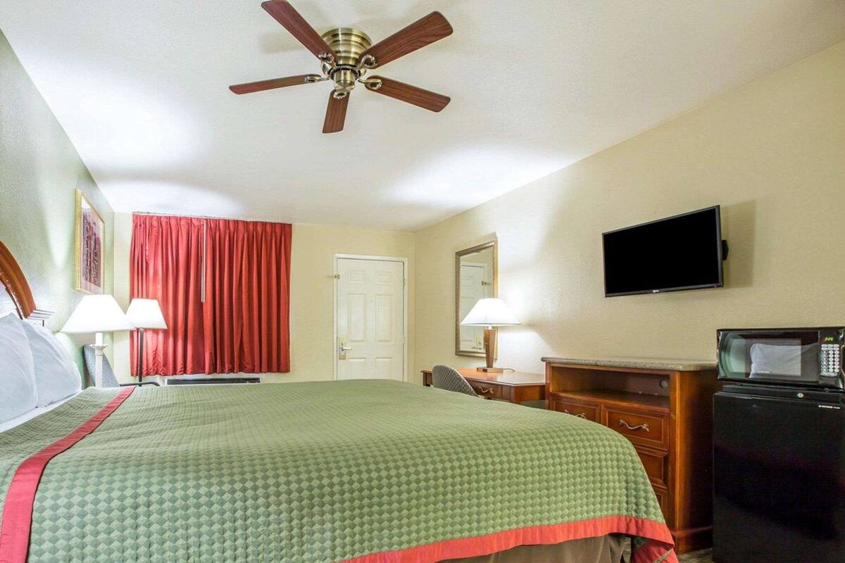 Quarto de hotel com cama, escrivaninha, televisão, micro-ondas e ventoinha de teto.