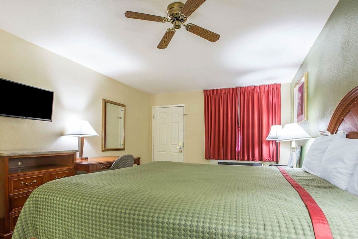 Quarto de hotel com cama, escrivaninha, cadeira, televisão, ventilador de teto e janela com cortinas.