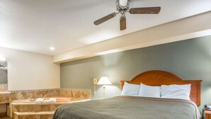 A hotel room with a bed, a ceiling fan, a bathroom with a bathtub, and a nightstand with a lamp.