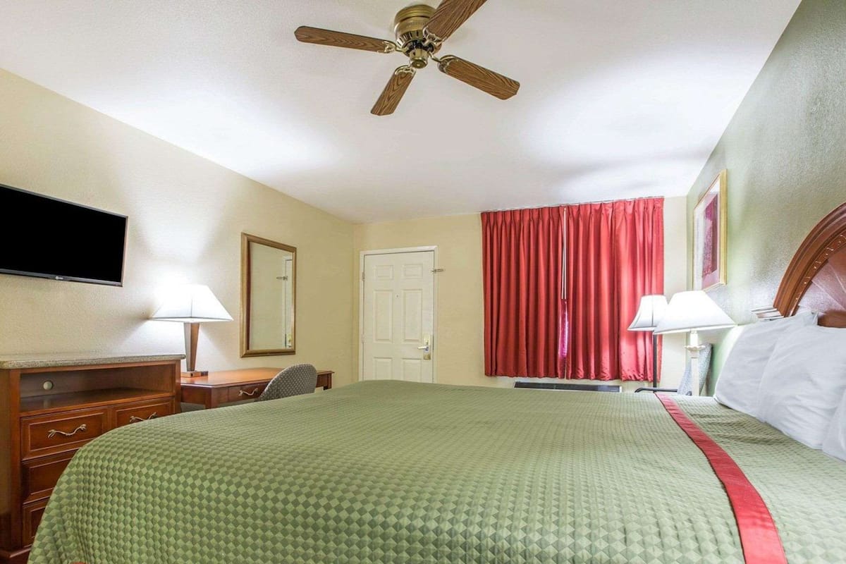 Quarto de hotel com cama, escrivaninha, cadeira, televisão, ventoinha no teto e janela com cortinas vermelhas.