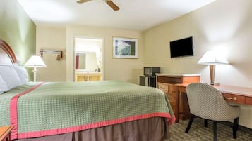 Habitación de hotel con cama, escritorio, silla, ventilador de techo, televisor y baño visible a través de una puerta abierta.
