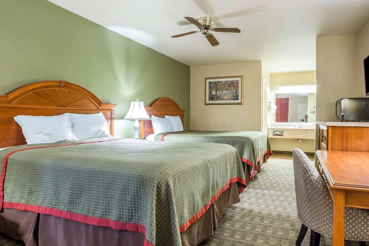 Quarto de hotel com duas camas, uma escrivaninha, uma cadeira, ventoinha no teto e um banheiro visível por uma porta aberta.