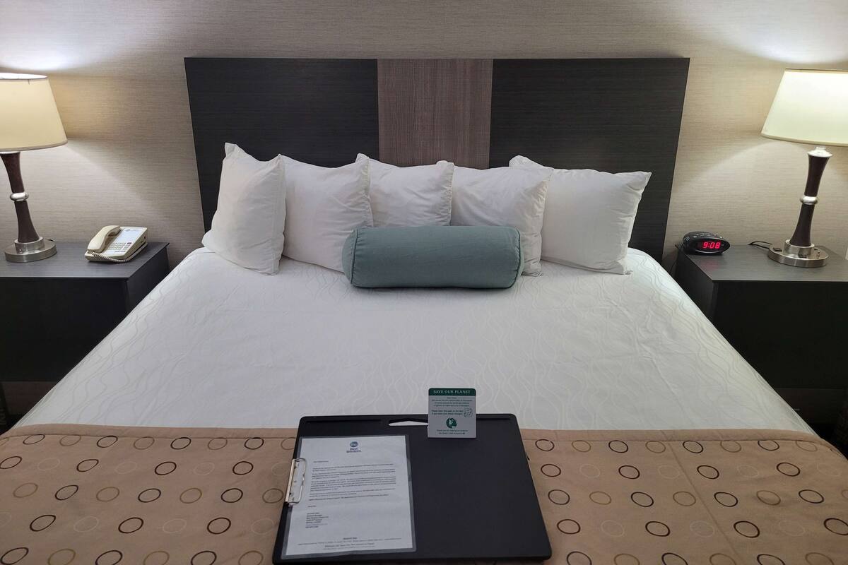 A hotel room with a neatly made bed, a bedside phone, a digital clock, and a tray with a document on the bed.