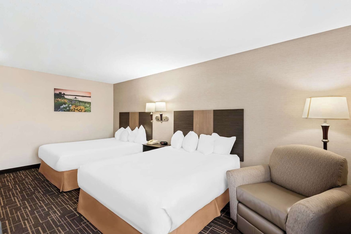 A hotel room with two beds, a desk, a chair, and a lamp.