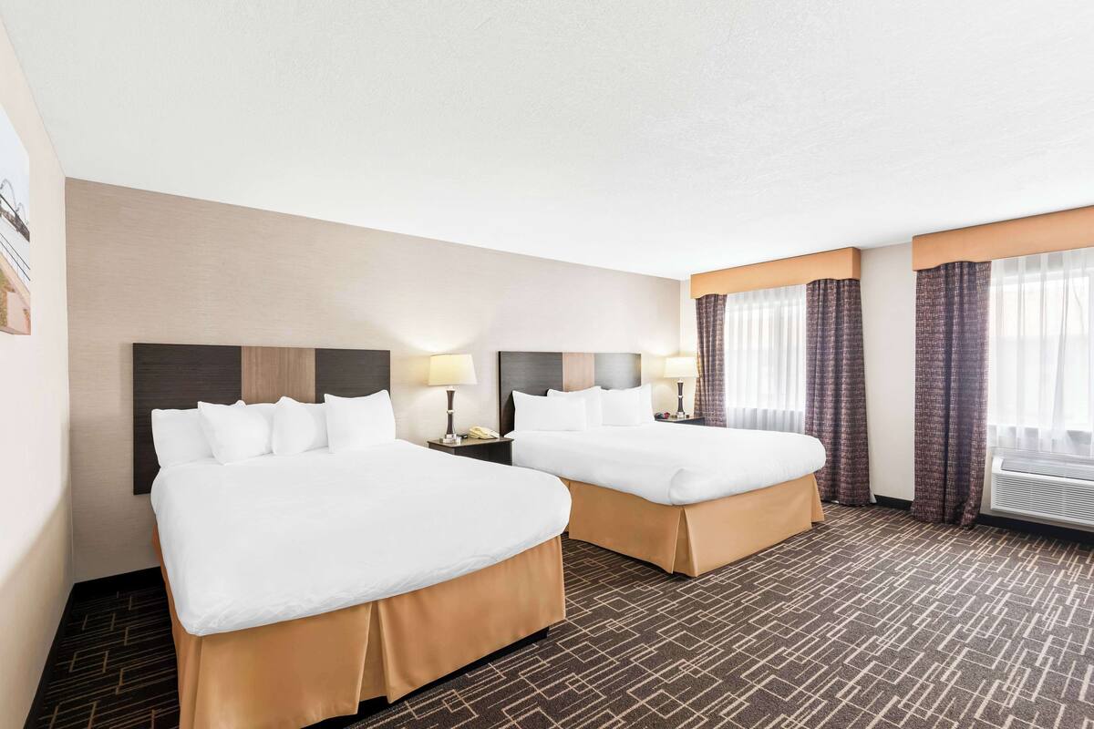 A hotel room with two beds, a large window with curtains, a wall-mounted air conditioning unit, and a patterned carpet.