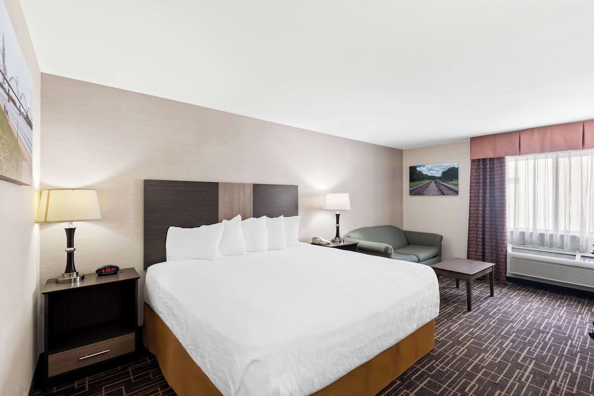 A hotel room with a large bed, two bedside tables with lamps, a small table, a green armchair, and a window with curtains.