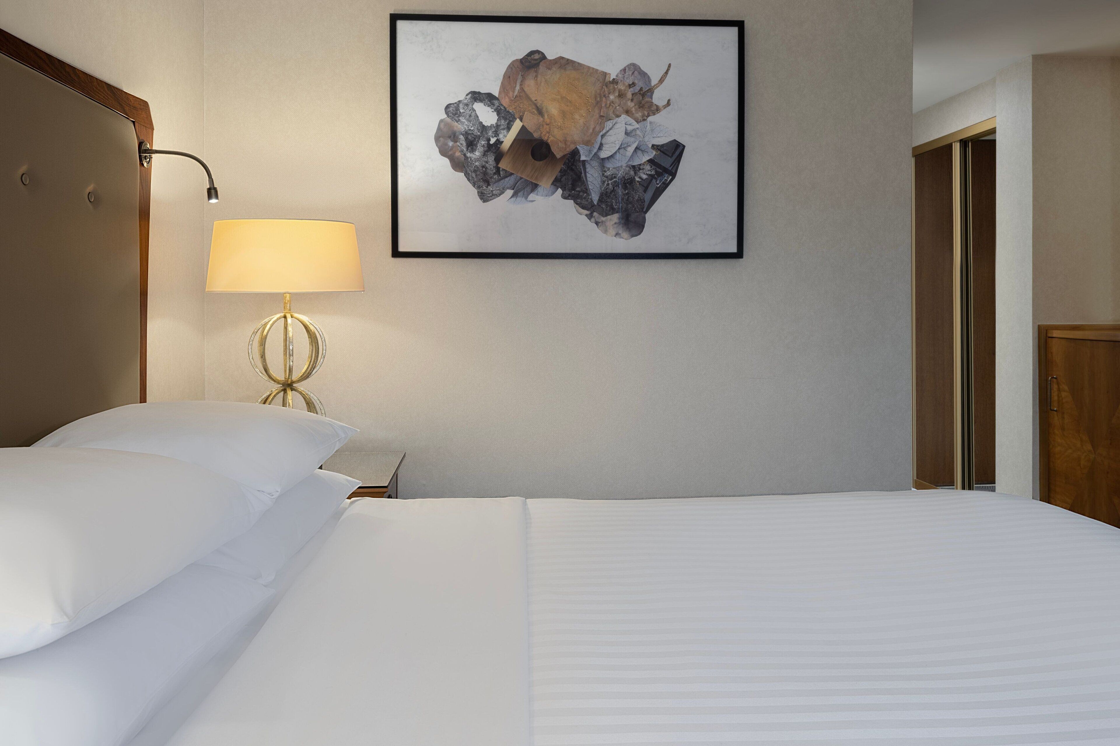Una habitación de hotel con una cama bien tendida, una lámpara y una obra de arte abstracta enmarcada en la pared.