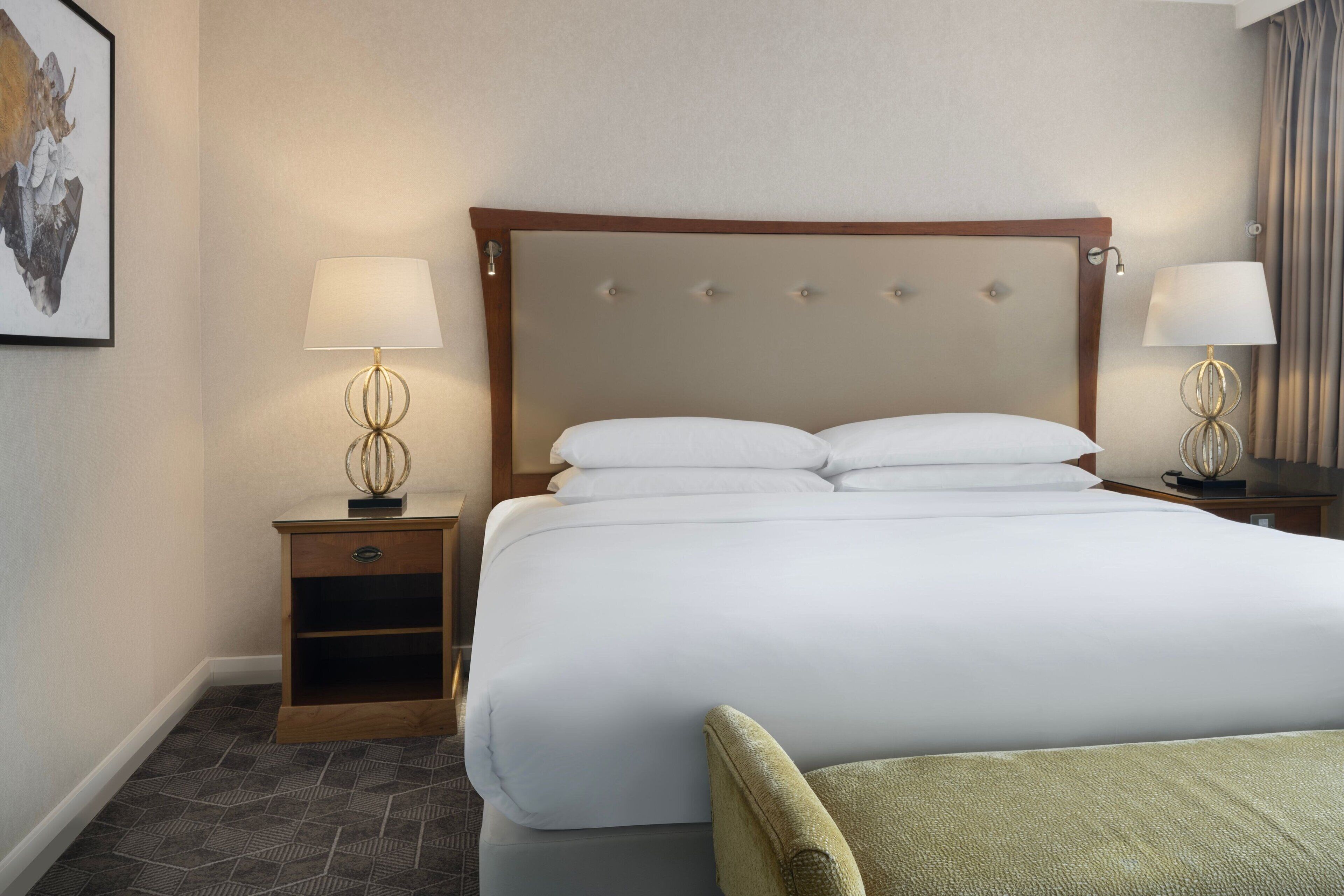 A neatly made bed with a wooden headboard, two bedside tables with lamps, a small drawer, and a patterned carpet.