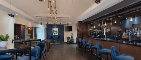 A bar area with wooden flooring, blue bar stools, hanging plants, and a bar counter with various items.