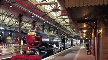 Una locomotora de vapor antigua en el andén de una estación de tren, con techo cubierto y cerca blanca.