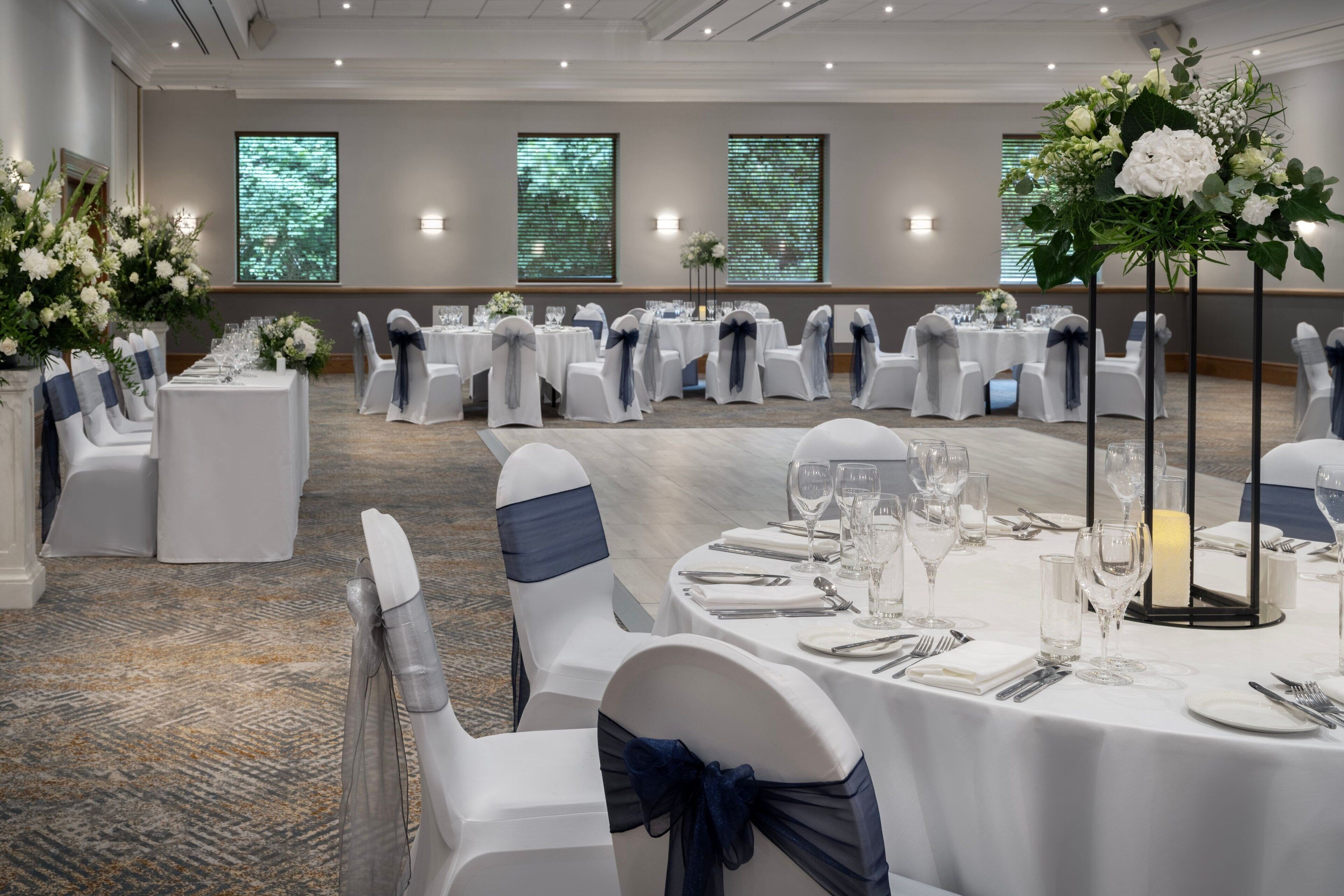 A banquet hall with round tables set for dining, white chairs with blue sashes, and floral centerpieces.
