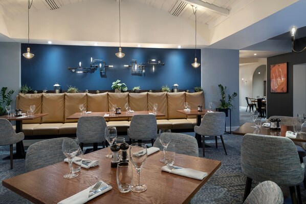 A restaurant with a blue wall, wooden tables, and cushioned seating. - Delta Hotels by Marriott Swindon (Swindon)