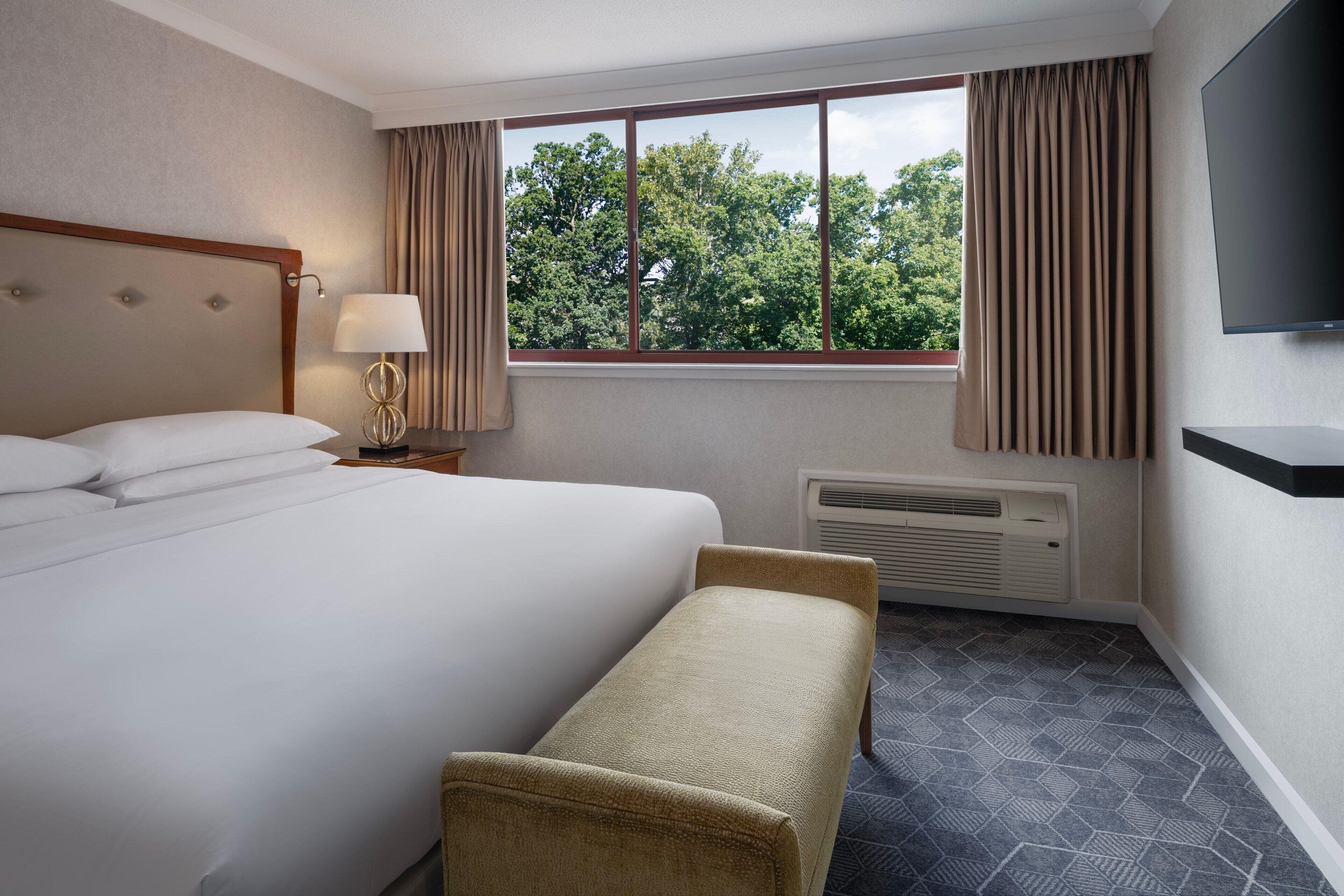 Een hotelkamer met een groot bed, een bankje, een televisie, een raam met gordijnen en een radiator.