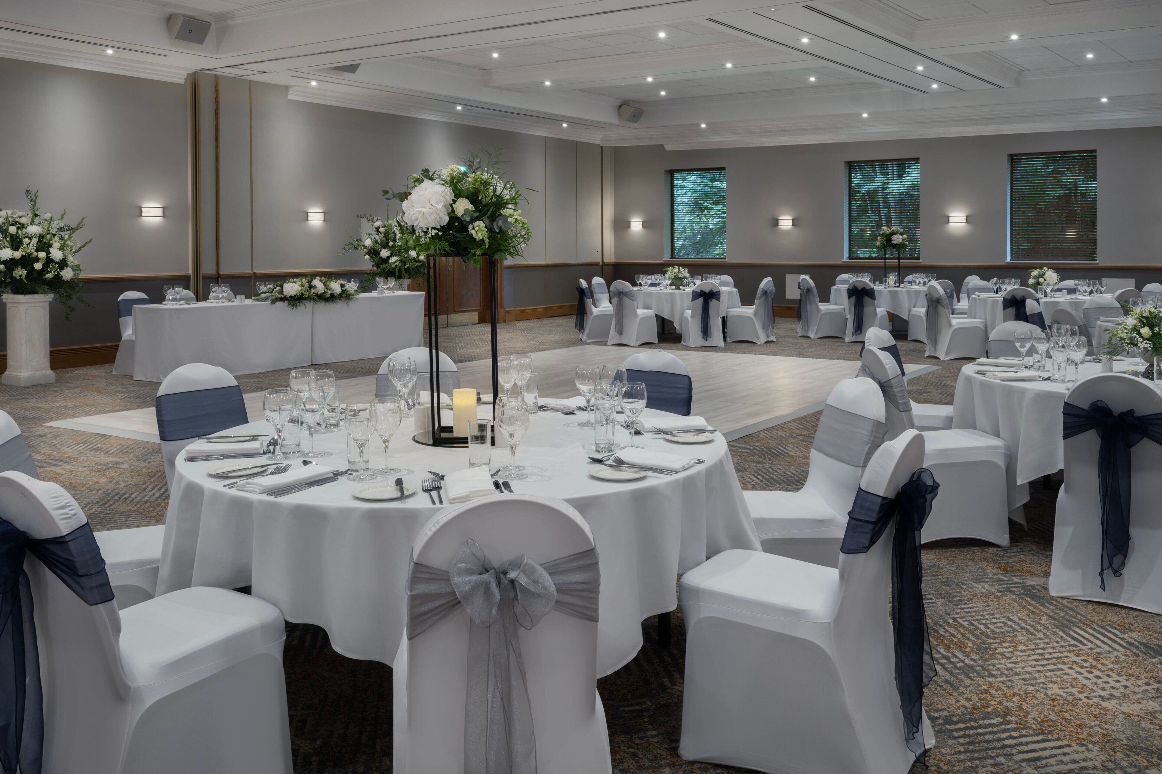 A banquet hall with round tables set for dining, white chairs with blue sashes, and floral centerpieces.
