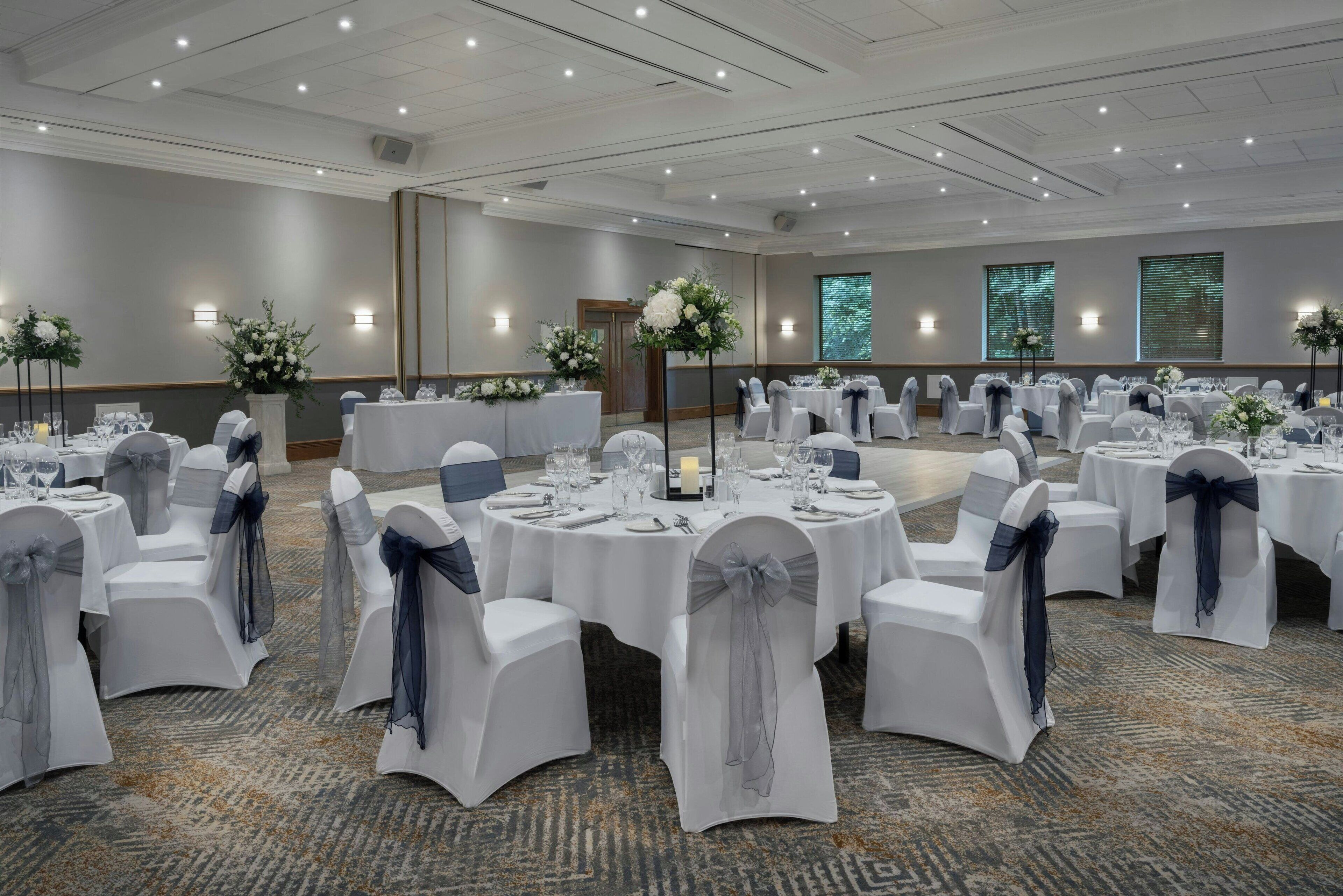 Une salle de banquet avec des tables rondes dressées pour des événements, des chaises blanches ornées de rubans bleus et des compositions florales en guise de centre de table.