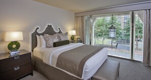 A hotel room with a large bed, two bedside tables with lamps, a bench, and a view of the outdoors through large windows.