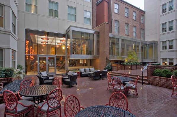 An outdoor patio area with seating and tables, a glass entrance, and a brick building in the background.
