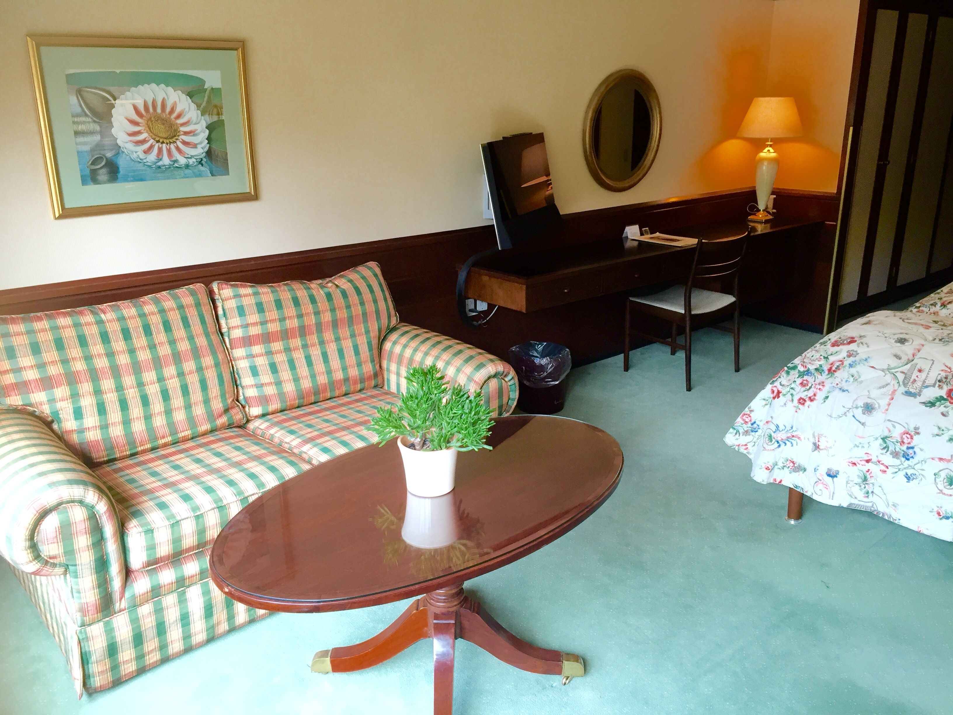 Habitación de hotel con sofá a cuadros, una pequeña mesa redonda con una planta, escritorio con lámpara, cama con ropa de cama floral y un cuadro en la pared.