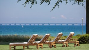 Four wooden lounge chairs with wheels on a grassy area, overlooking a body of water and a pier in the distance.