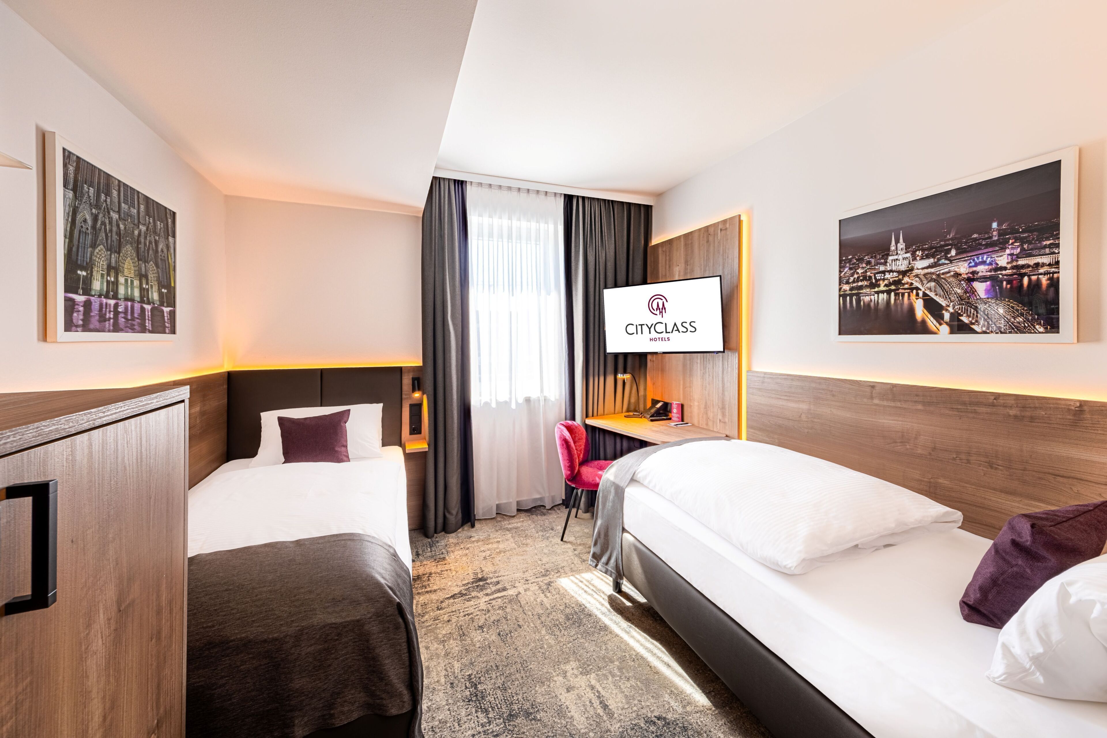 Ein Hotelzimmer mit einem Bett, einem Schreibtisch, einem Sessel und zwei Einzelbetten. An der Wand befindet sich ein Fernseher und ein Fenster mit Vorhängen.