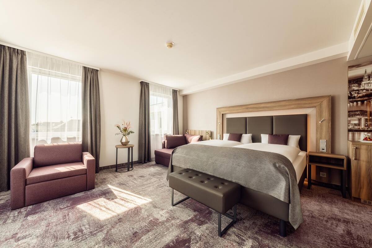 Une chambre d’hôtel avec un grand lit, un canapé, une banquette, une petite table avec un vase et une armoire.