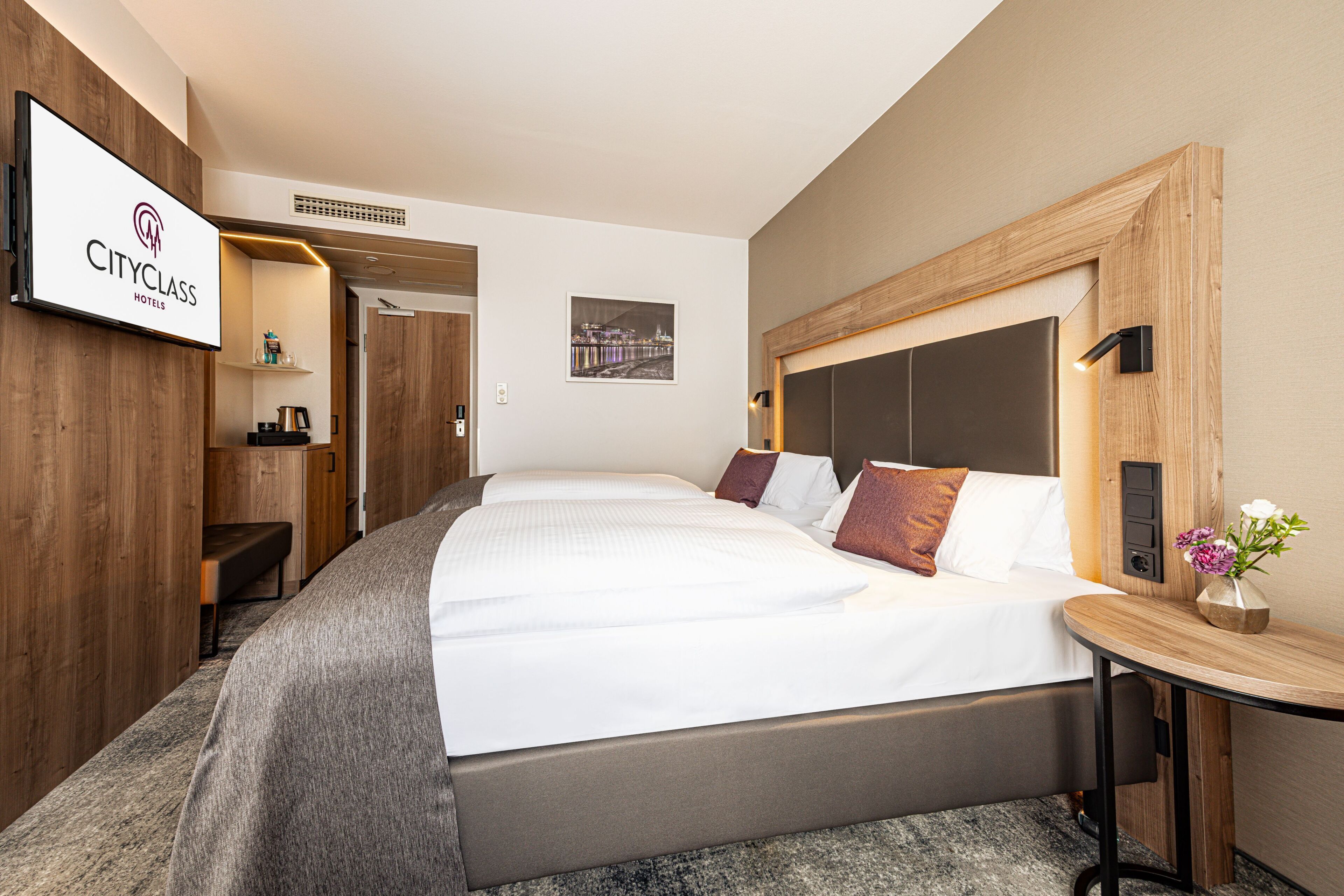 A hotel room with a large bed, a bedside table with a vase of flowers, and a wall-mounted TV displaying the hotel name.