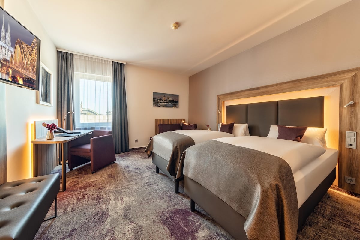 Une chambre d’hôtel avec deux lits, un bureau, une chaise et une grande fenêtre avec des rideaux.