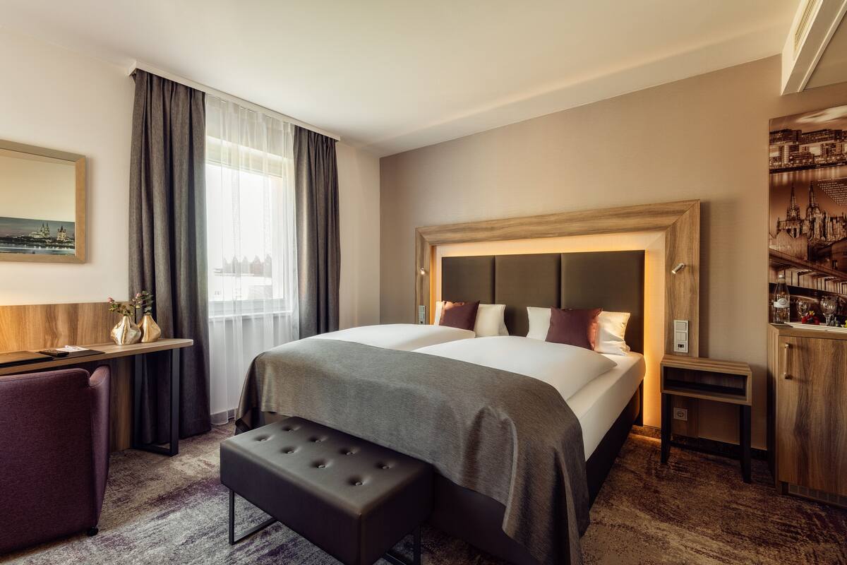 Une chambre d’hôtel avec un grand lit, un bureau, une chaise, un banc et une armoire.