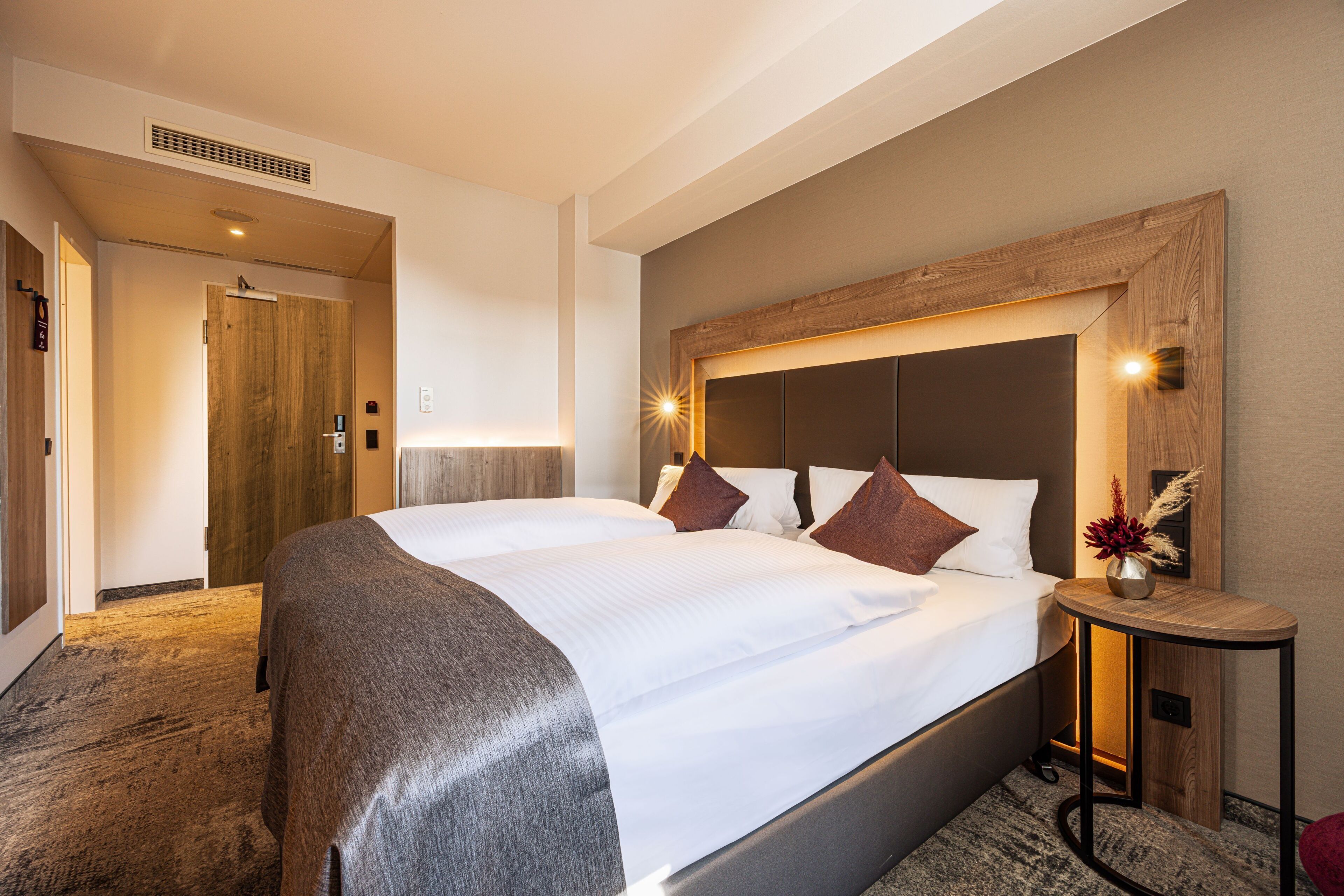 A hotel room with a large bed, a wooden headboard, a bedside table with a vase, and a carpeted floor.