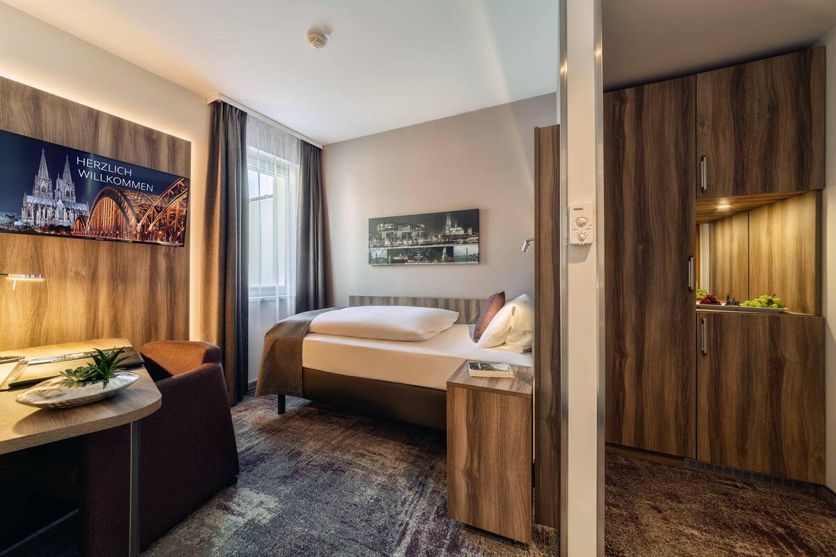 Une chambre d’hôtel avec un lit, un bureau, une chaise, une petite table avec une plante et un téléviseur fixé au mur qui diffuse des images d’un paysage urbain.
