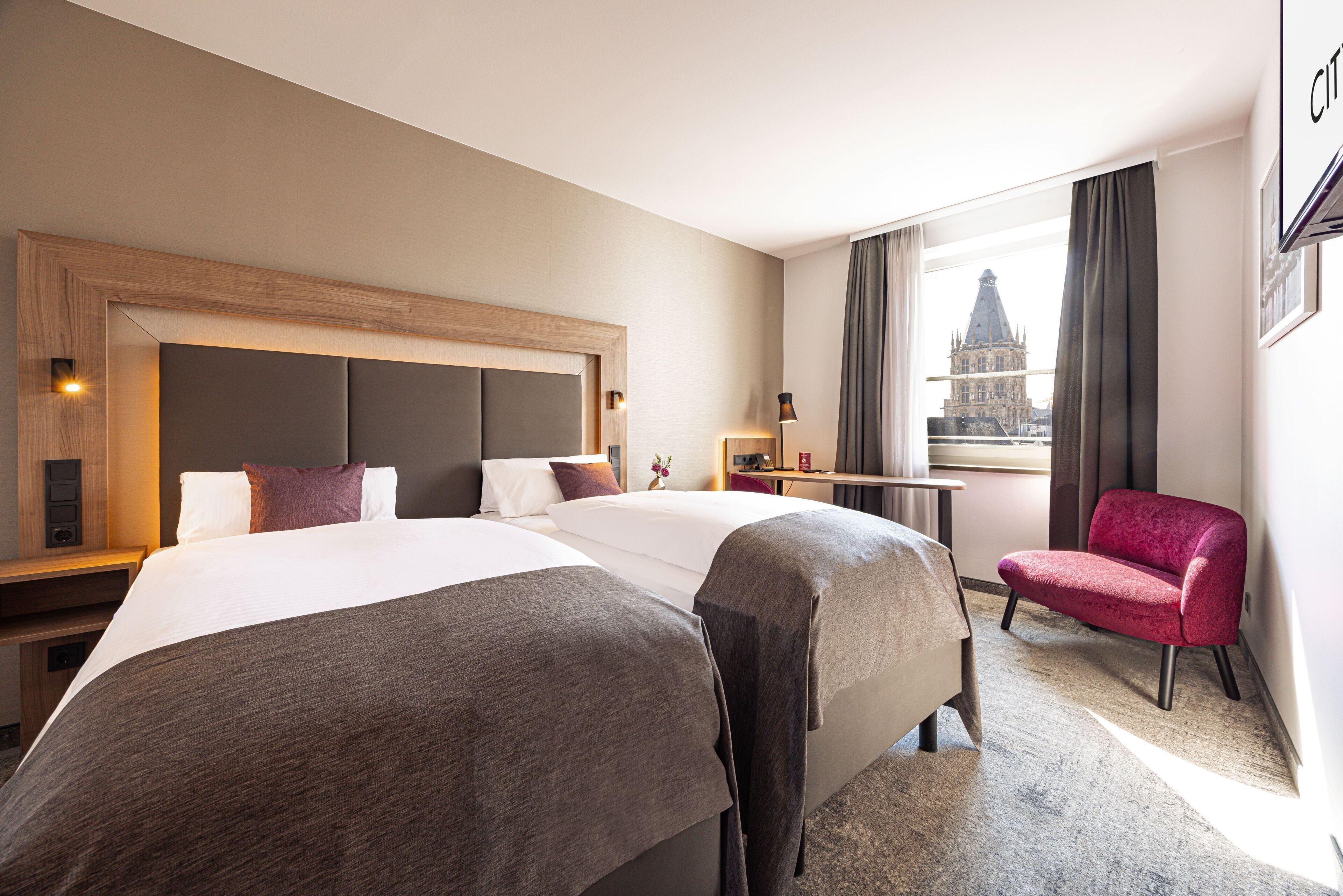 A hotel room with two beds, a desk, a chair, and a view of a building through the window.