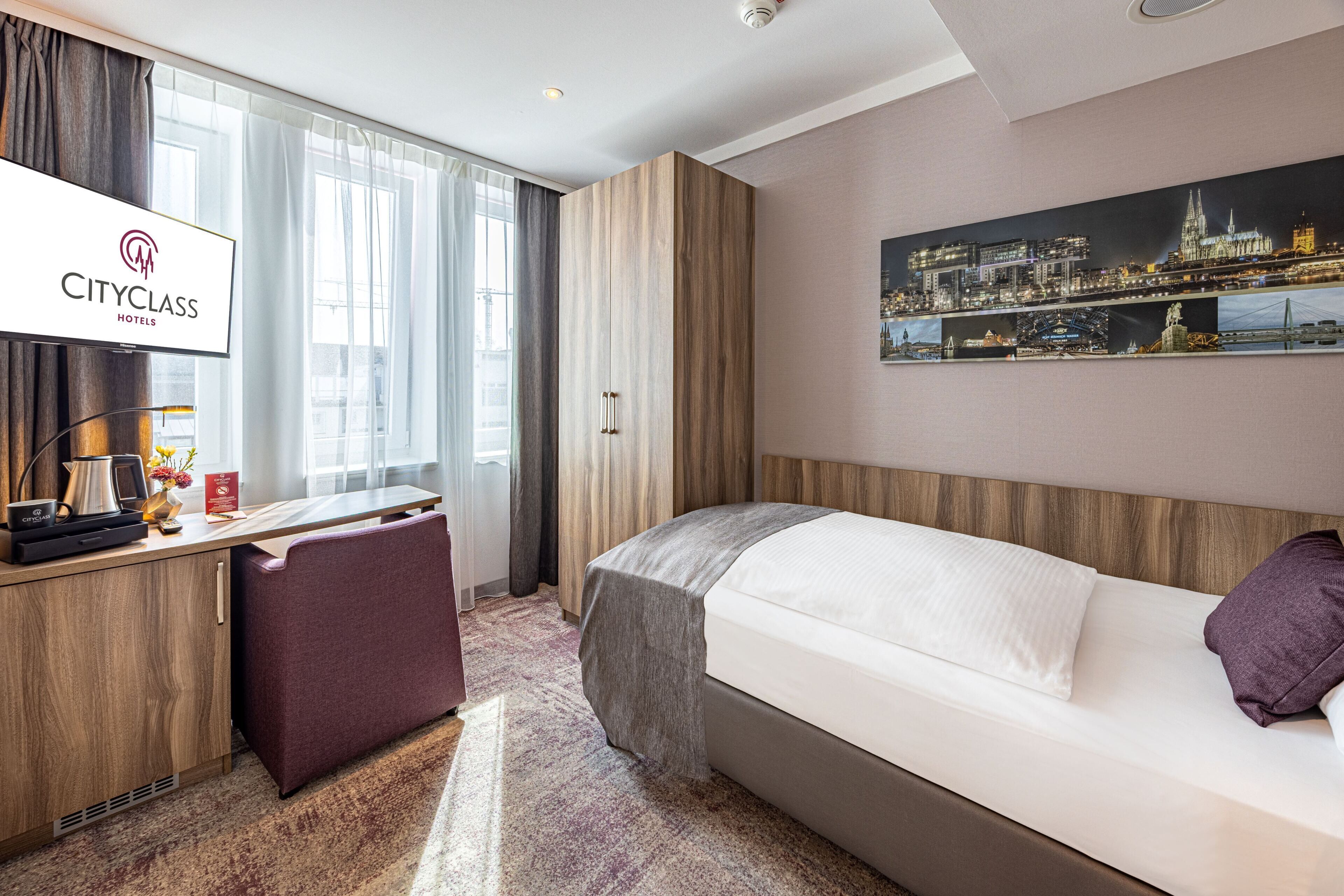 Ein Zimmer der CityClass Kategorie mit Bett, Schreibtisch, Stuhl, einer Kaffeemaschine und einem Stadtbild an der Wand.