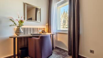 Habitación de hotel con escritorio, espejo, florero con plantas, ventana con cortinas y vistas a una catedral.
