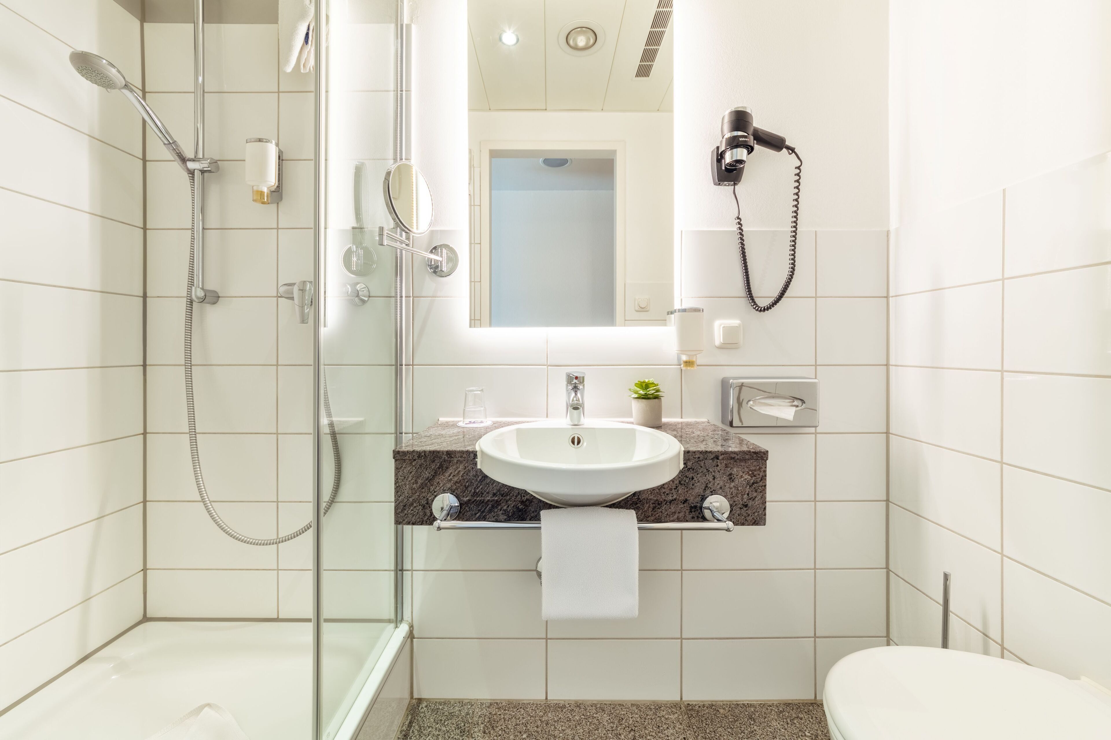 A modern bathroom with a glass-enclosed shower, a sink with a marble countertop, a hairdryer, and a toilet.