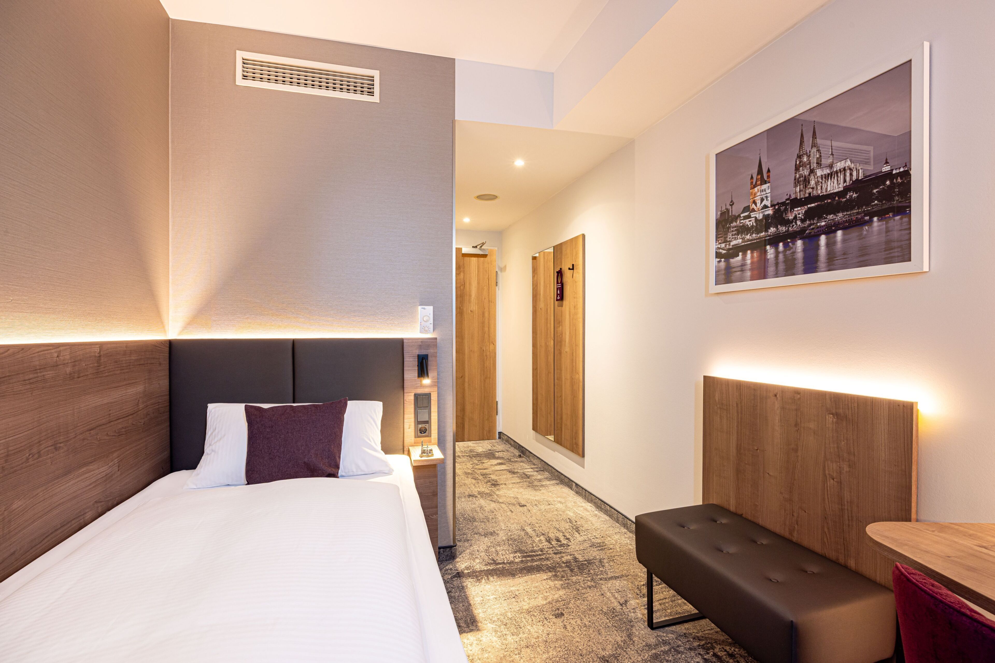 Een hotelkamer met een bed, nachtkastje, bankje en een stadslandschap schilderij.