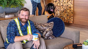A man sitting on a couch with a mug, a dog wearing a vest, and a receptionist behind a counter.