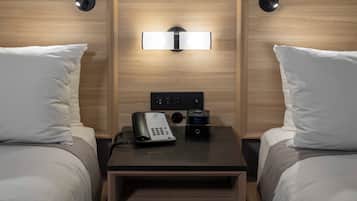 A hotel room with two beds, a bedside table with a phone and a digital clock, and a wall-mounted light fixture.
