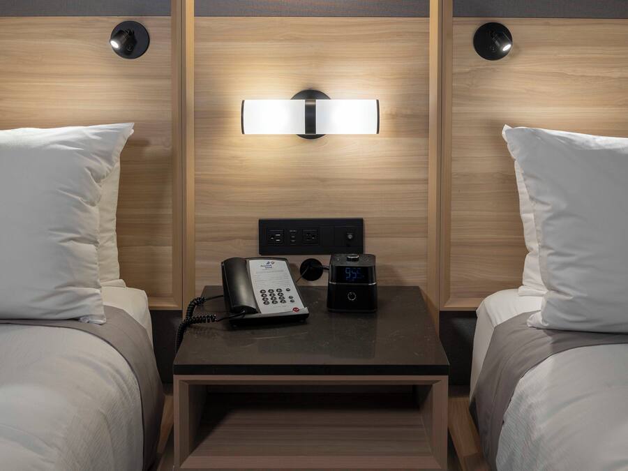 A hotel room with two beds, a bedside table with a phone and a digital clock, and a wall-mounted light fixture.