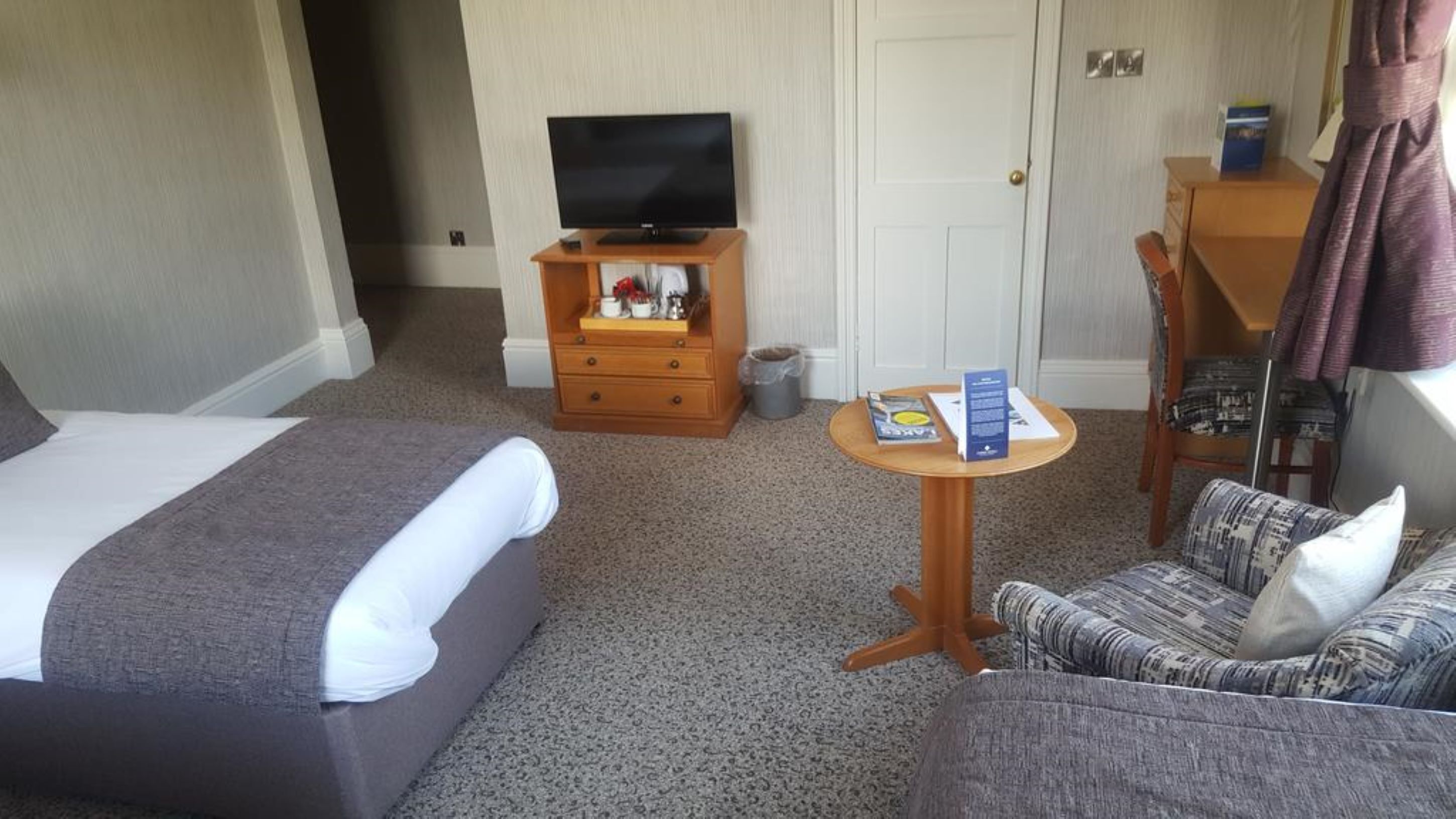 A hotel room with a bed, a sofa, a small table with a book, a television, and a door.