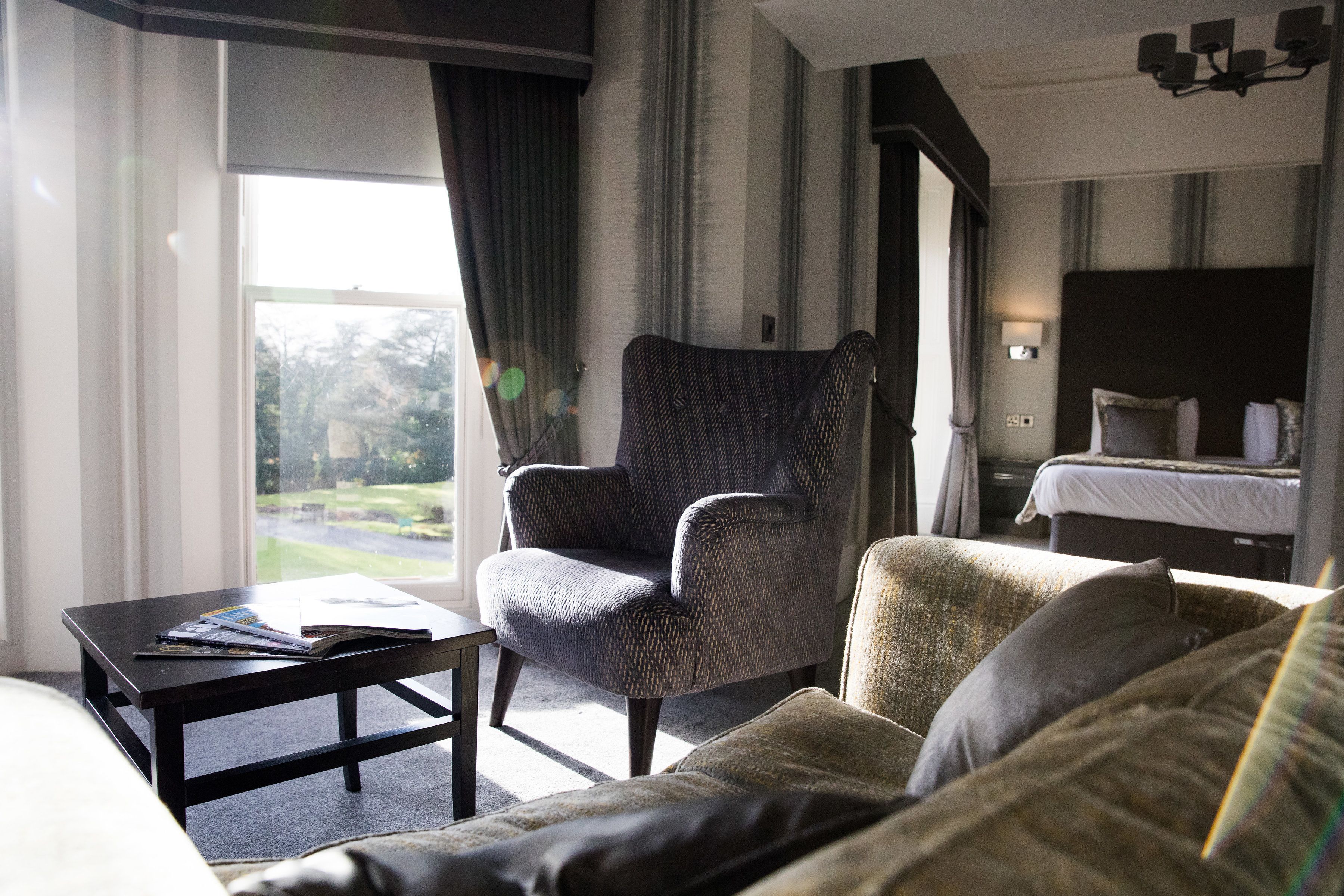 Une chambre d’hôtel avec une grande fenêtre, un fauteuil sombre, un canapé, une table de chevet et un lit avec une tête de lit.