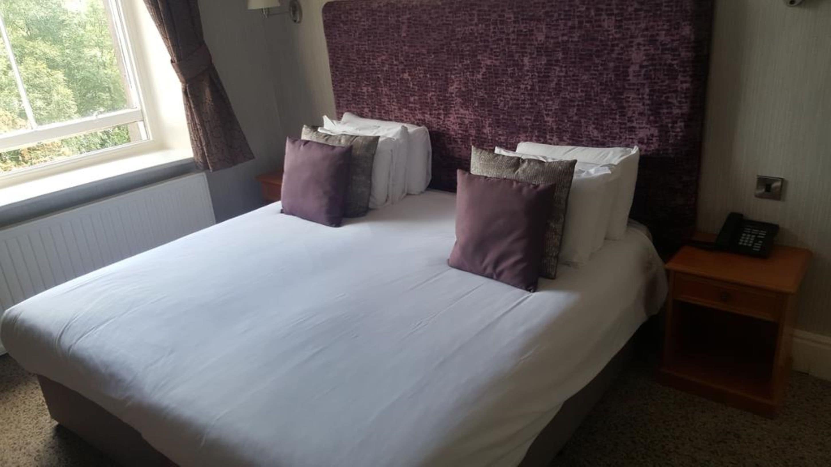 A neatly made bed with white linens and two purple pillows, a wooden nightstand with a phone, and a window with curtains.